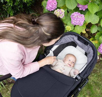 Mother smiling at baby lying comfortably in Joie Finiti Flex Travel Ready pram surrounded by garden flowers
