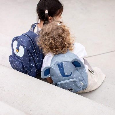 Children wearing backpacks sitting outdoors, one with a blue Backpack - Mrs. Elephant featuring ears and a trunk design, the other with a navy blue backpack with a swan motif